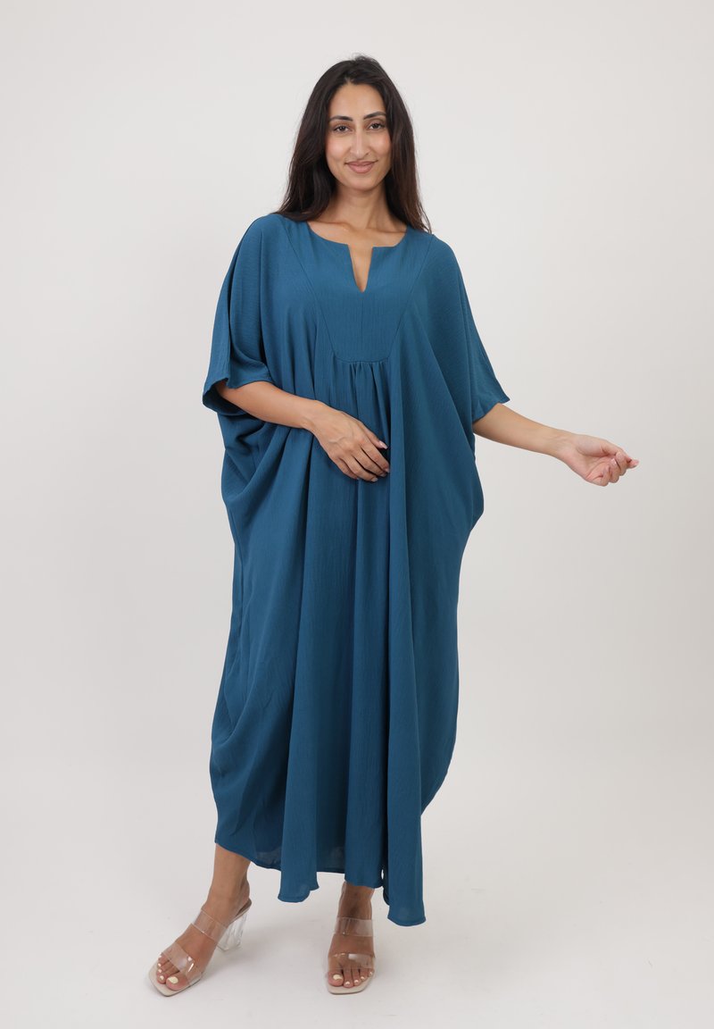 Kaftan turquoise, ample, avec des manches larges, un décolleté en V et un design fluide. Fabriqué en tissu léger au toucher lisse. Sandales à talons transparentes.
