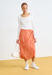 Long, wrap-around skirt in coral with a floral pattern; paired with a white ribbed long-sleeve shirt and white sneakers. Features a white handbag.