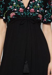 Schwarzes Kleid mit floraler Stickerei in Pink und Grün, V-Ausschnitt, gebundenem Taillenzug und einem Quasten-Detail am Saum.