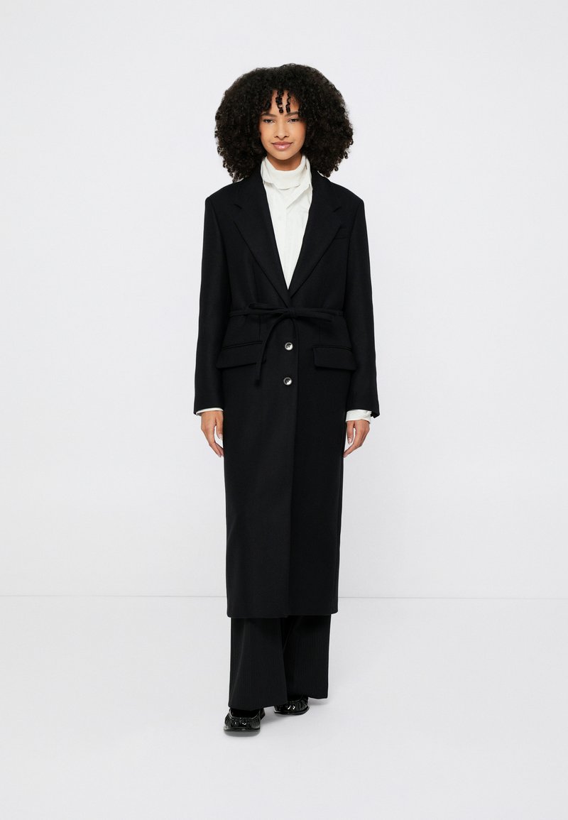 Must pikk mantel, millel on vöökohal rihm, kaks eesmine taskut ja suured nööbid. Kantud valge kraega särgi ja mustade laia säärega pükste peal.