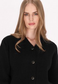 Schwarze Strickjacke mit V-Ausschnitt, ausgestattet mit großen metallischen Knöpfen und einem strukturierten Muster. Der Stoff wirkt weich und warm.