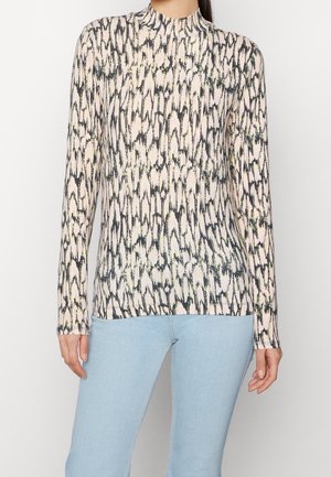 Femme portant un haut à manches longues et col montant avec un motif abstrait noir et beige, associé à un jean bleu clair.