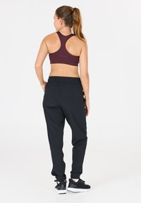 Bordeauxfarbener Sport-BH mit Racerback-Design, kombiniert mit schwarzen, taillierten Jogginghosen mit einem elastischen Bund, getragen mit schwarzen Sport-Schuhen.
