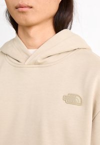 Sudadera beige hecha de un tejido suave con un bolsillo tipo canguro en la parte delantera. Presenta un logo bordado tonal en el pecho izquierdo y una capucha grande.