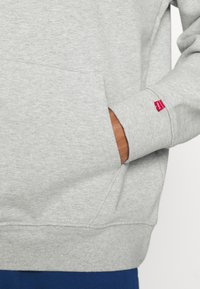 Felpa grigia in pile con tasca a marsupio, polsini a costine e una piccola etichetta rossa Levi's sulla manica. Tessuto morbido, design casual.