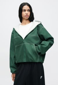 Veste verte zippée avec capuche grise, présentant des détails de coutures blanches et des poignets élastiques. Portée sur une chemise blanche et associée à un pantalon noir.