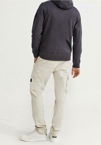Sudadera gris con capucha y bolsillo canguro, combinada con pantalones cargo beige claros con bolsillos laterales, y zapatillas blancas con detalles en negro.