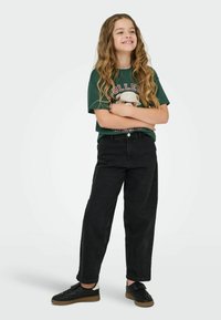 Zelené grafické tričko s designem medvěda, spárované s volnými černými džínovými kalhotami a černými teniskami s hnědými podrážkami. Dlouhé, vlnité vlasy.