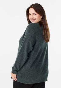 Tamsiai žalia megzta palaidinė, laisvo kirpimo, su ilgomis rankovėmis ir šiek tiek siaurėjančiu apačia. Faktūrinis audinys su matomais megztumo raštais.