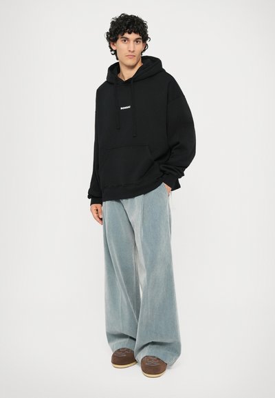 Sweat-shirt à capuche noir avec une poche avant et un logo blanc, associé à un pantalon large bleu clair et des chaussures marron.