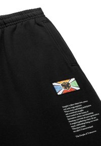 Zwarte joggingsbroeken hebben een kleurrijke patch met een ontwerp van een zwarte roos op een achtergrond van gekleurde segmenten. Er is tekst op het been gedrukt.