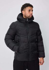 Veste matelassée noire avec capuche, dotée d'une fermeture éclair à l'avant, de deux poches latérales zippées, et d'un design à panneaux cousus pour l'isolation.