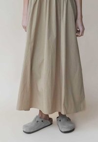 Sabots en suede gris avec une forme arrondie, dotés d'une sangle ajustable et d'une semelle en caoutchouc noir, associés à une longue jupe beige.