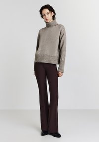 Pull à col roulé gris clair avec un tricot texturé, associé à un pantalon foncé et ajusté marron. Le pull présente un ourlet et des poignets côtelés.
