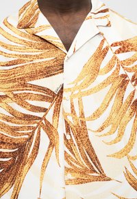 Άνδρας που φοράει ένα λευκό πουκάμισο με κουμπιά, με πορτοκαλί και καφέ τροπικό φύλλωμα και φαρδιά γιακά.