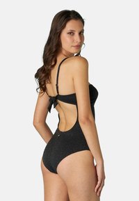 Costume da bagno intero nero in tessuto testurizzato, con spalline regolabili, schiena scoperta e un dettaglio a fiocco in vita. Presenti sottili accenti metallici.