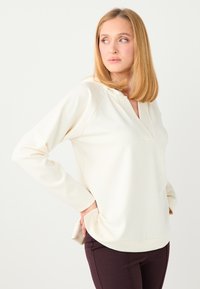 Haut à manches longues de couleur crème avec un col en V et des manches raglan, présentant une texture lisse et des poches latérales, associé à un pantalon marron foncé.