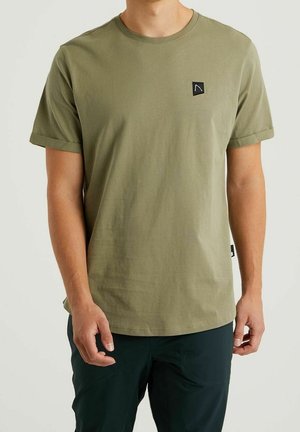 Man draagt een olijfgroen T-shirt met korte mouwen en een klein zwart logo op de borst, gecombineerd met donkere broek, staand tegen een effen achtergrond.