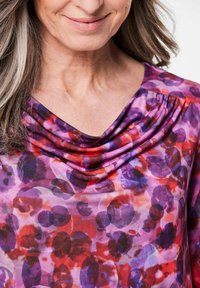 Lila Bluse mit einem drapierten Ausschnitt, versehen mit einem aquarellartigen Muster aus roten, pinken und blauen kreisförmigen Formen auf einem hellen Hintergrund.