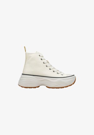 Zapatillas altas de lona blanca con una suela texturizada con patrón de ondas. Presenta ojales plateados, cordones blancos y una franja negra en contraste.