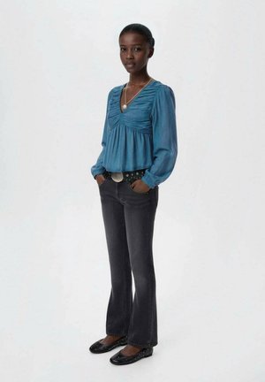Blusa blu a maniche lunghe con dettagli arricciati, abbinata a jeans neri a zampa d'elefante e scarpe flat, completata da una cintura decorata e un accento in argento.