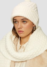 Witte gebreide beanie met een geribbeld ontwerp en een pompon, gecombineerd met een dikke, gestructureerde witte sjaal. Achtergrond is neutraal grijs.
