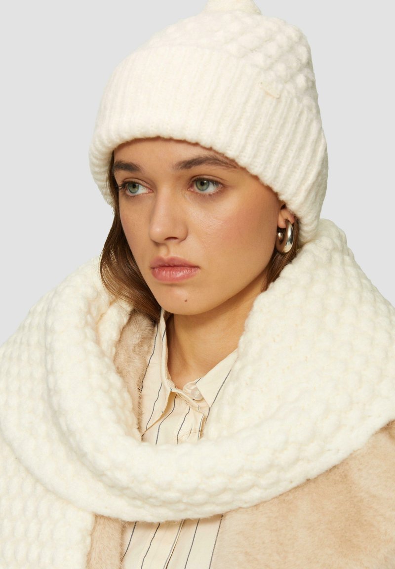 Witte gebreide beanie met een geribbeld ontwerp en een pompon, gecombineerd met een dikke, gestructureerde witte sjaal. Achtergrond is neutraal grijs.