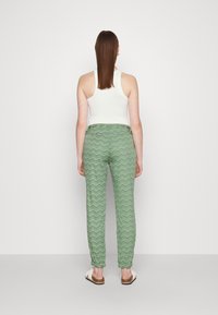 Woman standing facing away wearing a white sleeveless top, green zigzag patterned pants, and white slip-on sandals against a plain background.