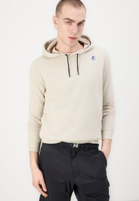 Hoodie beige chiaro con cordino e dettaglio del logo. Indossato con pantaloni cargo neri caratterizzati da molteplici tasche. Texture liscia, vestibilità morbida.