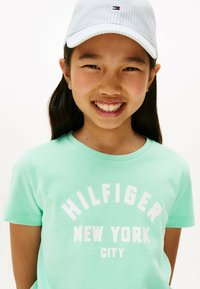 Camiseta de algodón verde menta con impresión gráfica blanca de "HILFIGER" y "NEW YORK CITY". Gorra de rayas azul claro con detalles del logo.