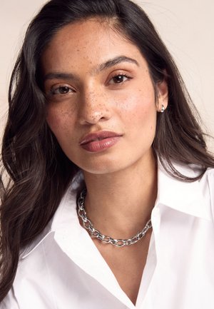 Silver chain necklace worn over a white collared shirt. The skin features freckles, and the hair is dark and wavy. Natural makeup.