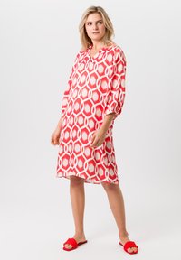 Rood en wit patroon jurk met geometrische vormen, losse pasvorm, V-hals en driekwartmouwen. Gecombineerd met rode slide sandalen.