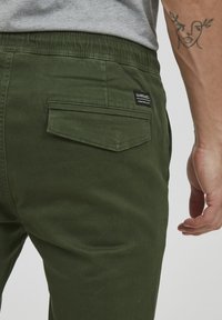 Green cotton pants with a textured finish, featuring a back pocket and an elastic waistband. Includes a small label near the pocket.