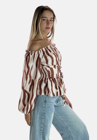Biała bluzka z dekoltem odsłaniającym ramiona, ozdobiona falującymi, rdzawymi paskami, z bufiastymi rękawami, marszczoną talią i baskinką. Połączona z jasnoniebieskim dżinsem.