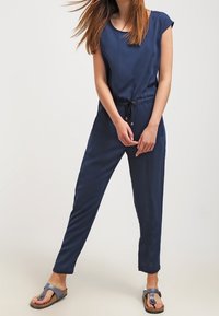 Femme portant une combinaison sans manches bleu marine avec une taille à cordon et des sandales grises à enfiler, se tenant devant un fond clair.