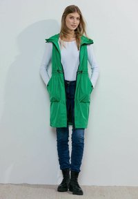 Gilet smanicato verde in piumino con cappuccio, dotato di tasche frontali, vita regolabile e chiusura con zip, indossato sopra una maglietta bianca a maniche lunghe e jeans blu.