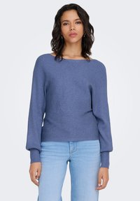 Pull en maille bleu foncé avec texture côtelée, col large et manches ballon. Associé à un jean bleu clair.