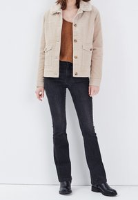 Femme portant une veste en velours côtelé beige, un haut en tricot marron, un jean noir ajusté et des bottines noires, posant devant un fond blanc.