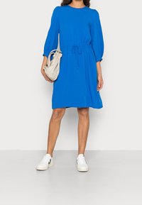 Sininen mekko, jossa on löysä muotoilu, kolme neljännesosaa hihat ja kiristettävä vyötärö. Yhdistetty valkoisiin urheilukenkiin ja vaaleanharmaaseen käsilaukkuun.