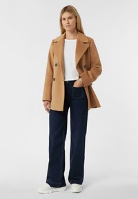 Cappotto cammello doppiopetto con rever a punta, indossato sopra una camicia bianca e jeans a gamba larga blu scuro; abbinato a sneakers bianche.