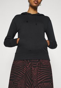 Sweat à capuche noir en tissu doux, doté d'une poche kangourou, d'une capuche réglable avec cordon de serrage et de poignets côtelés. Porté avec une jupe à motif.