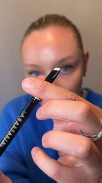 Person holding a black eyebrow pencil with a white tip close to the camera, wearing blue clothing and silver rings on fingers.