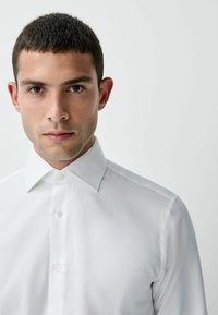 Valkoinen pitkähihaisessa paidassa on klassinen kaulus, nappikiinnitys ja sileä kangas, jossa on hienovaraisia ompeluyksityiskohtia ja hyvin istuva malli.