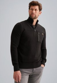 Schwarzer Pullover aus strukturiertem Stoff mit gerippten Details an den Ärmeln, kurzem Reißverschluss auf der Vorderseite und einer Brusttasche mit Reißverschluss.