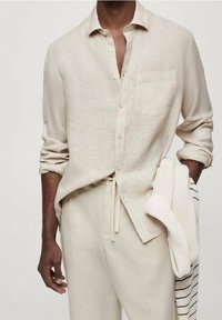 Beżowa lniana koszula z kołnierzykiem, z przodu na guziki, z jedną kieszenią na piersi i podwiniętymi rękawami. W połączeniu z beżowymi spodenkami na sznurek i prążkowanym swetrem.