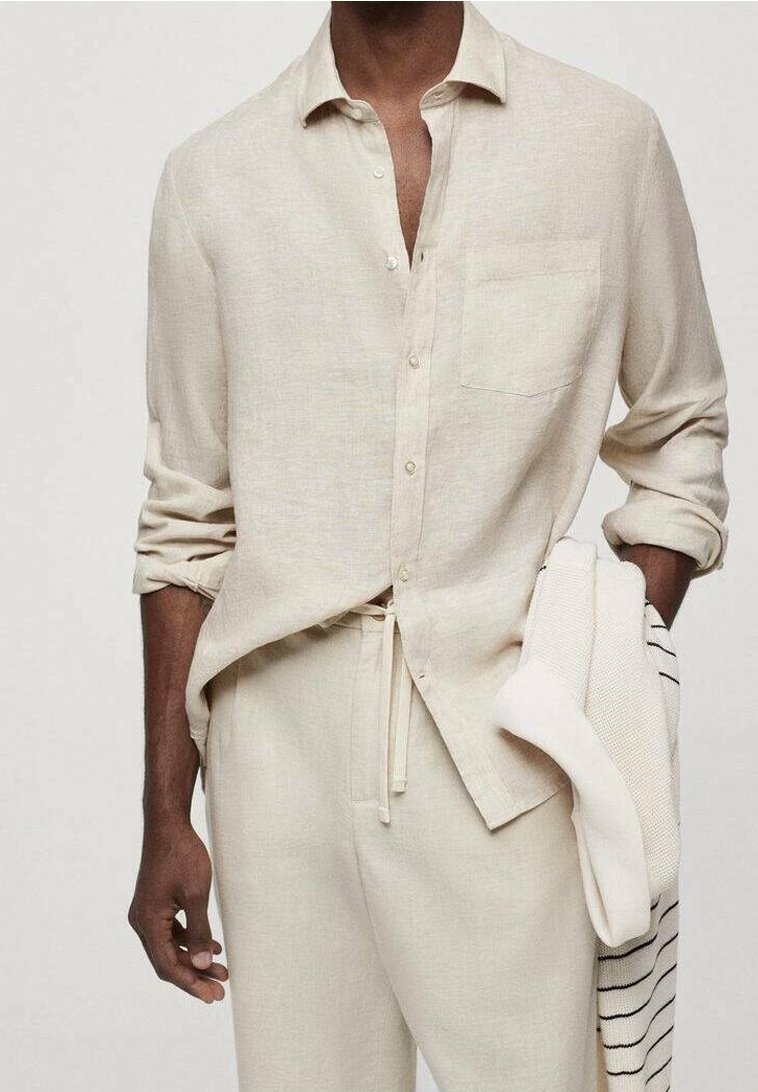 Beżowa lniana koszula z kołnierzykiem, z przodu na guziki, z jedną kieszenią na piersi i podwiniętymi rękawami. W połączeniu z beżowymi spodenkami na sznurek i prążkowanym swetrem.
