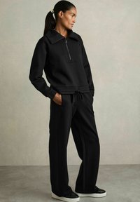 Felpa nera con colletto alto e mezza zip. Abbinata a pantaloni neri larghi e sneakers bianche. Tessuto liscio e morbido.