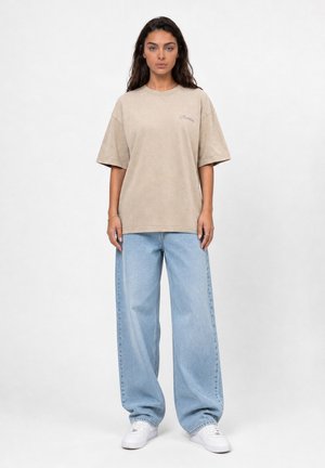 Vrouw staand met een beige oversized t-shirt, lichtblauwe wijde jeans en witte sneakers op een effen witte achtergrond.