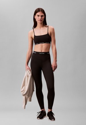 Kobieta ubrana w czarny sportowy stanik i legginsy Calvin Klein, trzymająca beżową bluzę, stojąca na szarym tle, ubrana w czarne trampki.