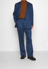 Completo in raso blu con giacca doppiopetto e pantaloni a gamba larga, abbinato a un dolcevita marrone a coste e scarpe nere.